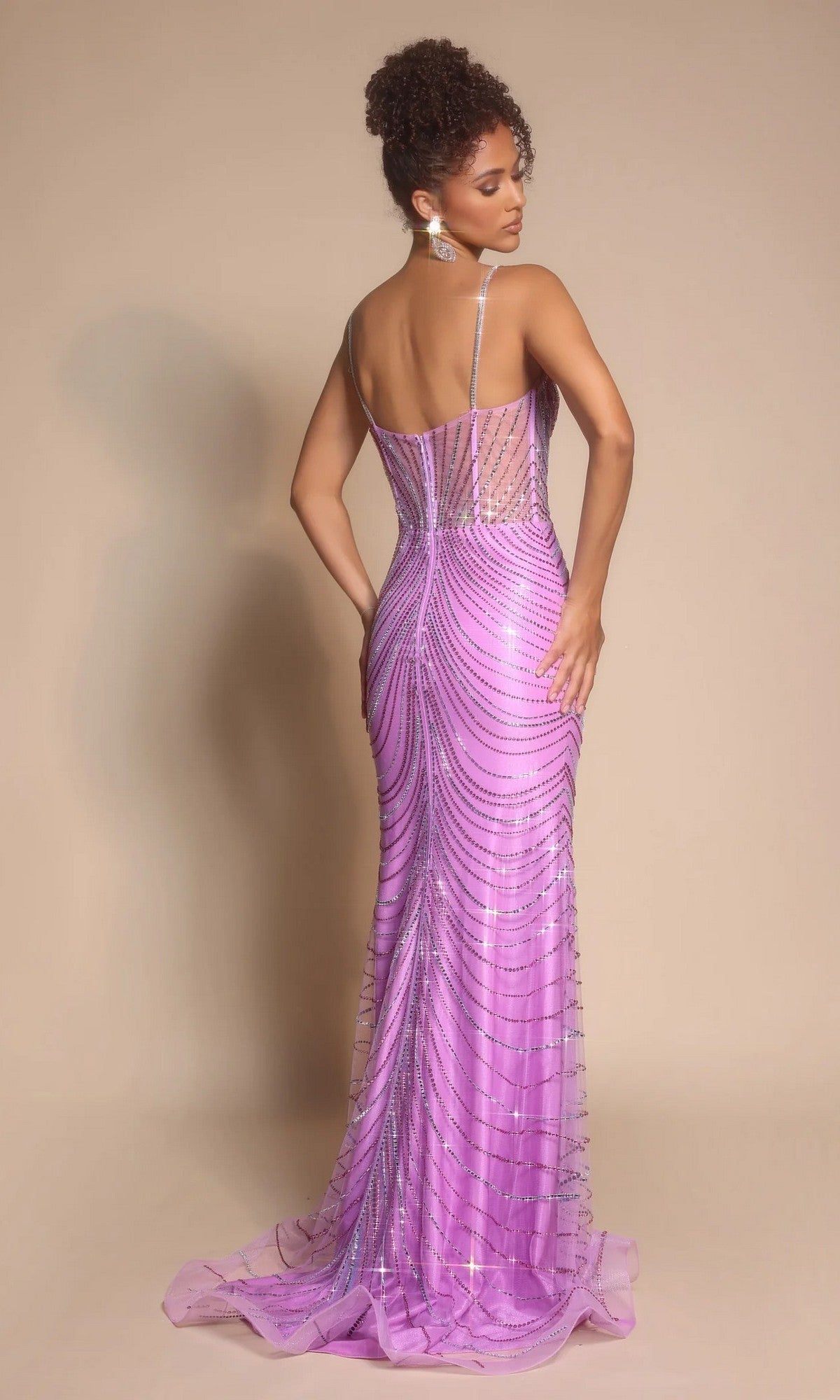 Woman in a fitted, floor-length Portia and Scarlett PS26095 lilac gown with thin straps and beaded details stands against a plain beige background, facing away from the camera.